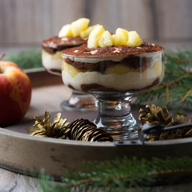 Tiramisu mit Bratapfel in einem hohen Dessertschälchen geschichtet mit Kakao und Apfelstückchen garniert. 